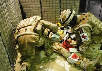 Soldiers in desert combat uniform, one with a red cross on his arm, treat a casualty on the ground